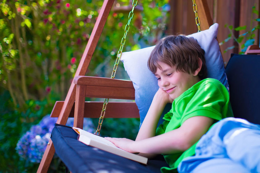 54639036 - happy school boy reading a book in the backyard. child relaxing in a garden swing with books. kids read during summer vacation. children studying. teenager kid doing homework outdoors.